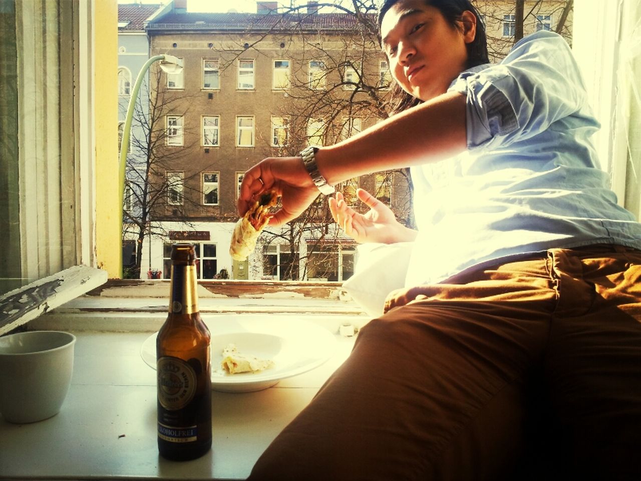 Portrait of man sitting on window sill and holding food by beer bottle
