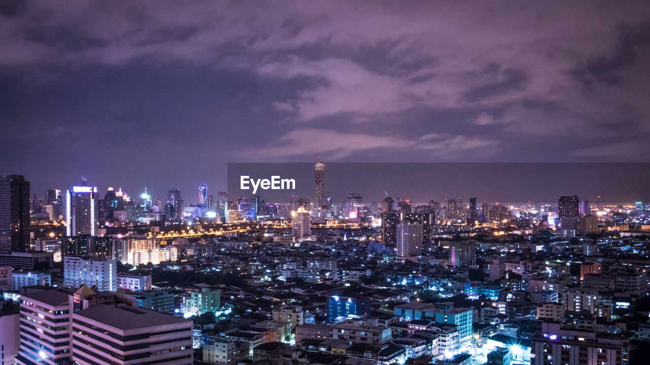 High angle view of illuminated cityscape against cloudy sky at night