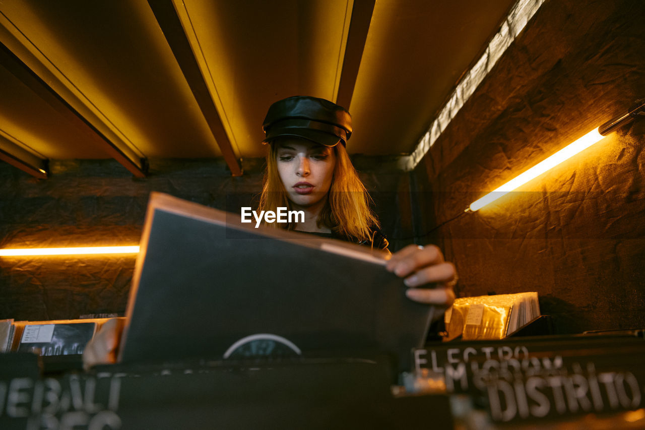 Front view of serene young female in black cap choosing vinyl record while standing near counter with collection of musical albums in store with glowing lamps