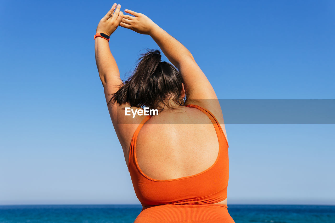 Back view of anonymous plump female athlete in sportswear exercising with raised arms against ocean under blue sky