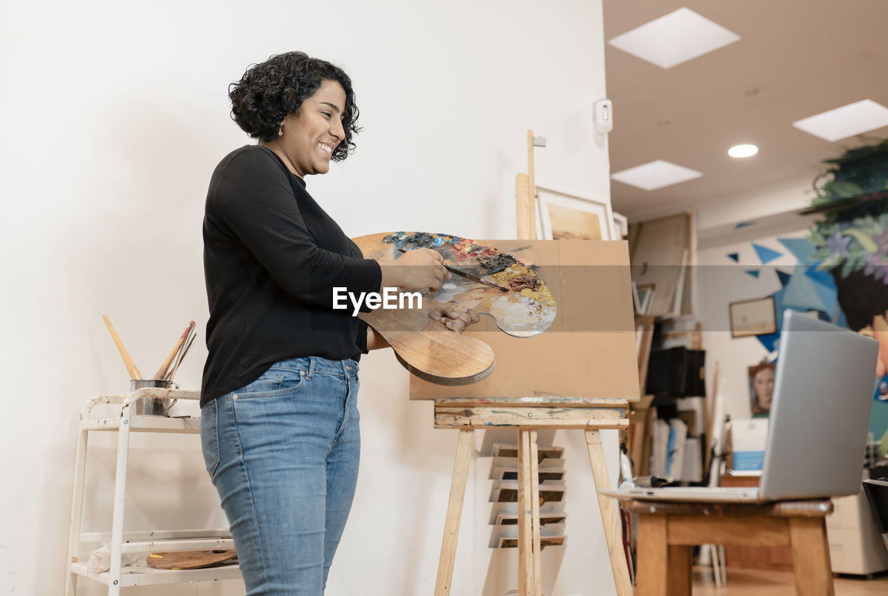 Smiling female artist on video call through laptop holding palette at studio