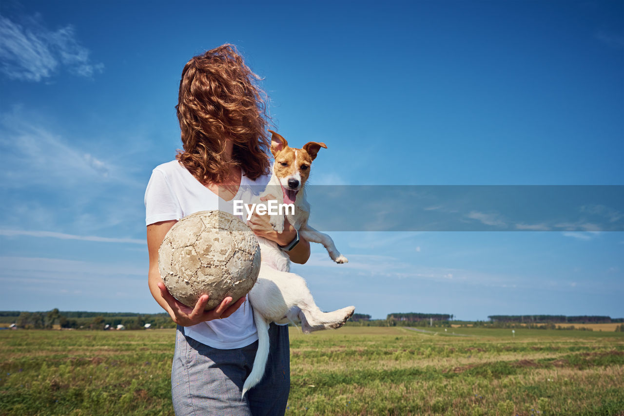 Woman holding dog with ball on field against sky