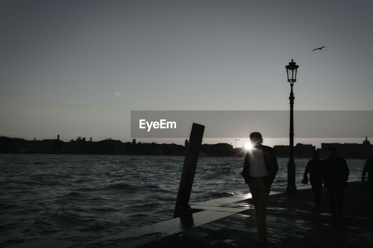 WOMAN STANDING BY SEA AGAINST SKY DURING SUNSET