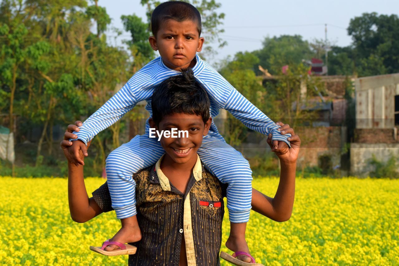 Portrait of smiling teenage boy carrying brother on shoulders at farm