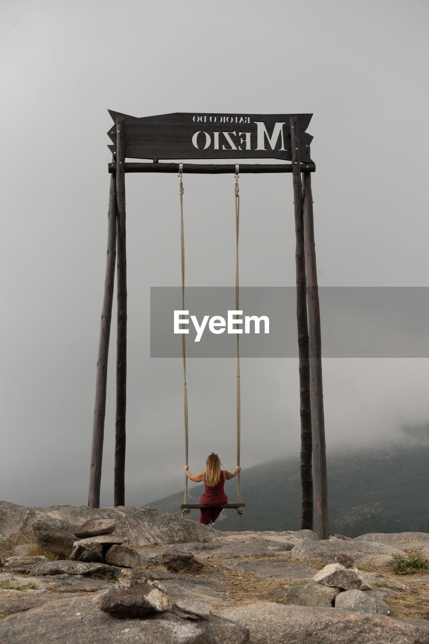 Rear view of woman sitting on rope swing against sky