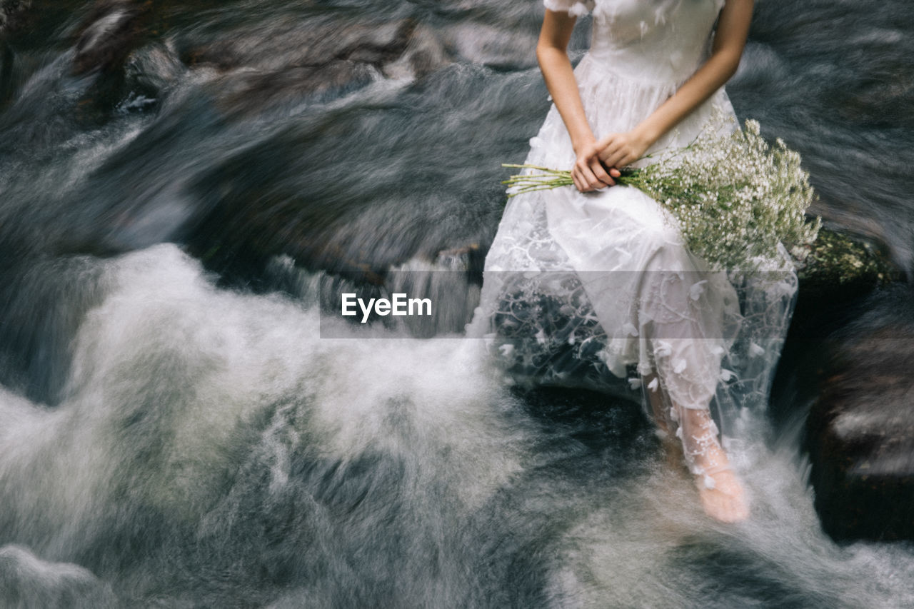 Low section of woman in wedding dress sitting on rock over river