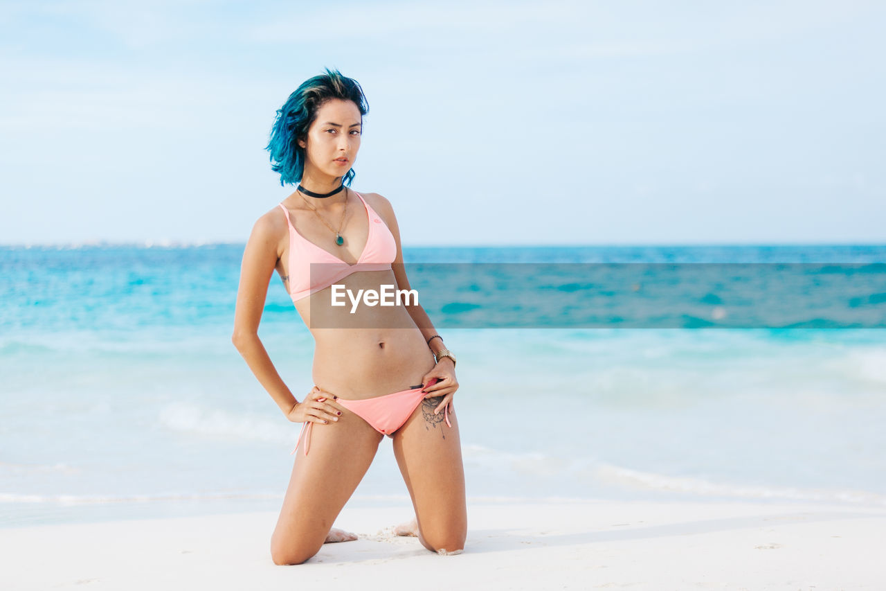 Portrait of woman in bikini kneeling on sand at beach against sky