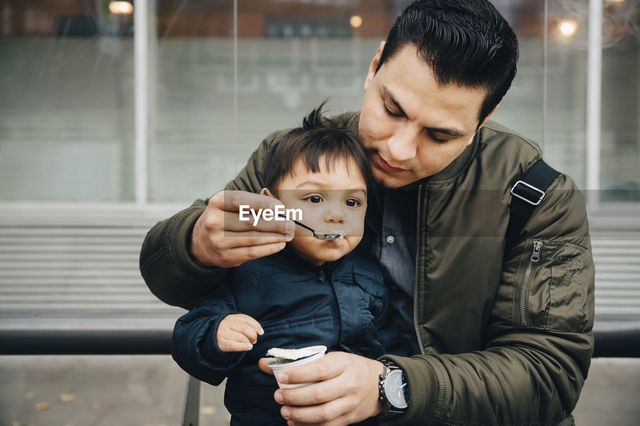 Father feeding baby food to son while sitting on bench in city