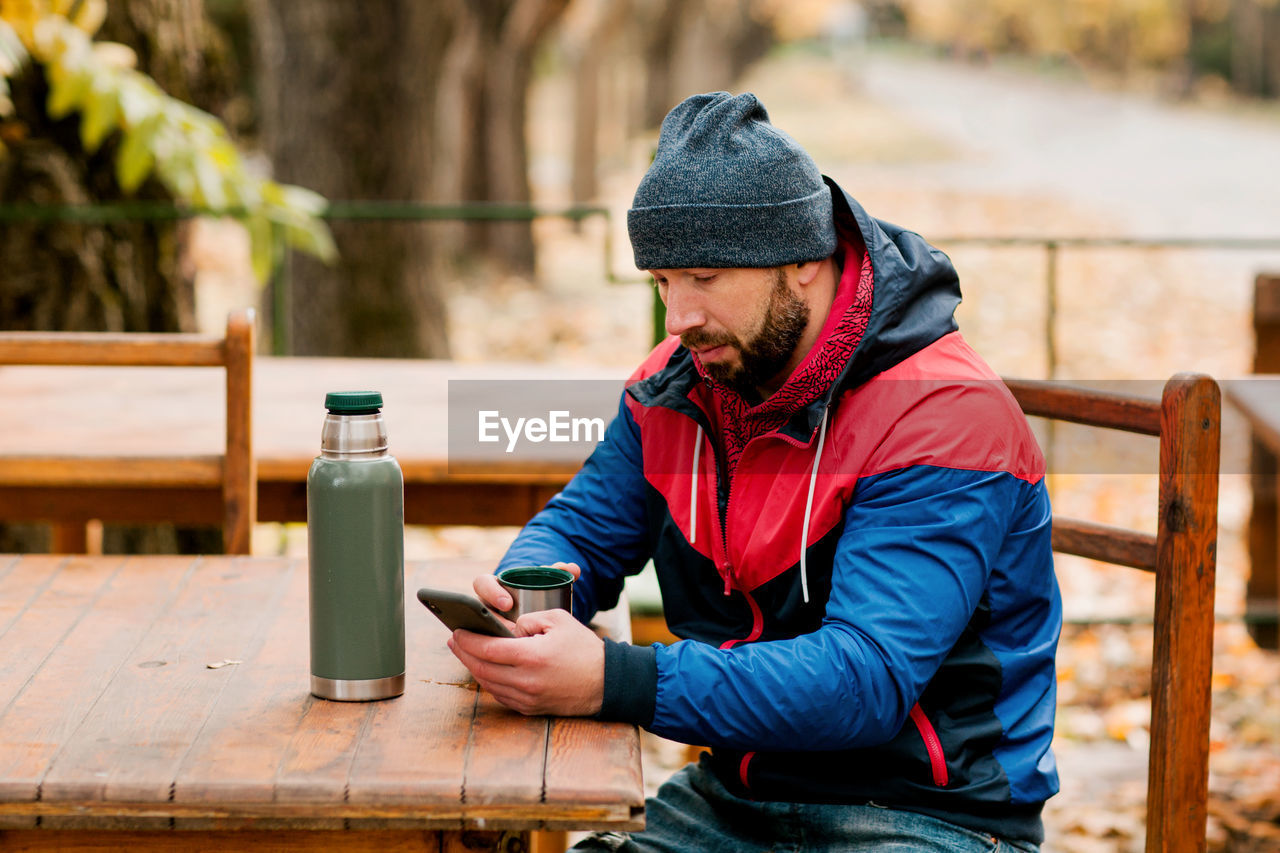 A young man using a mobile phone is sitting outdoors in a cafe or restaurant on an autumn day
