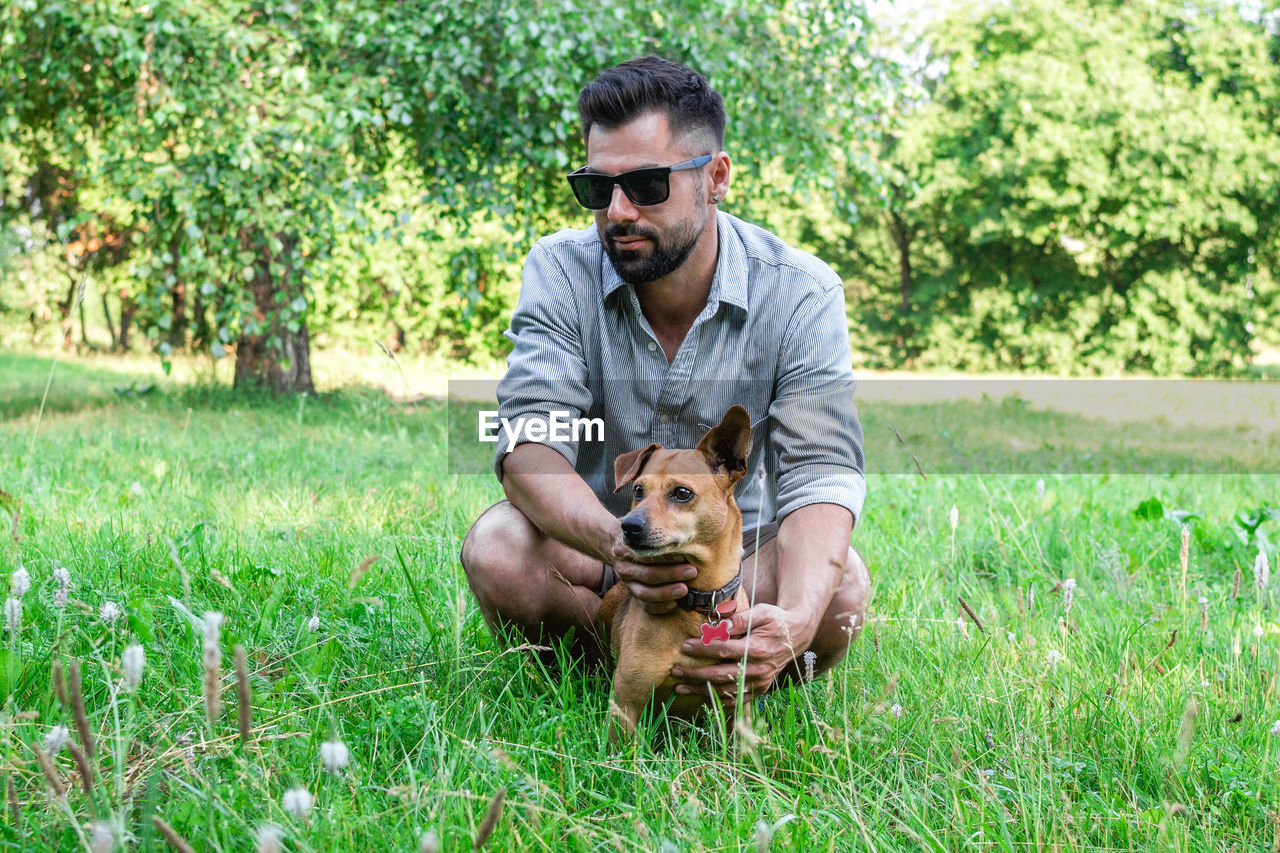 Handsome stylish european man sitting on grass with his dog in park on a walk. 