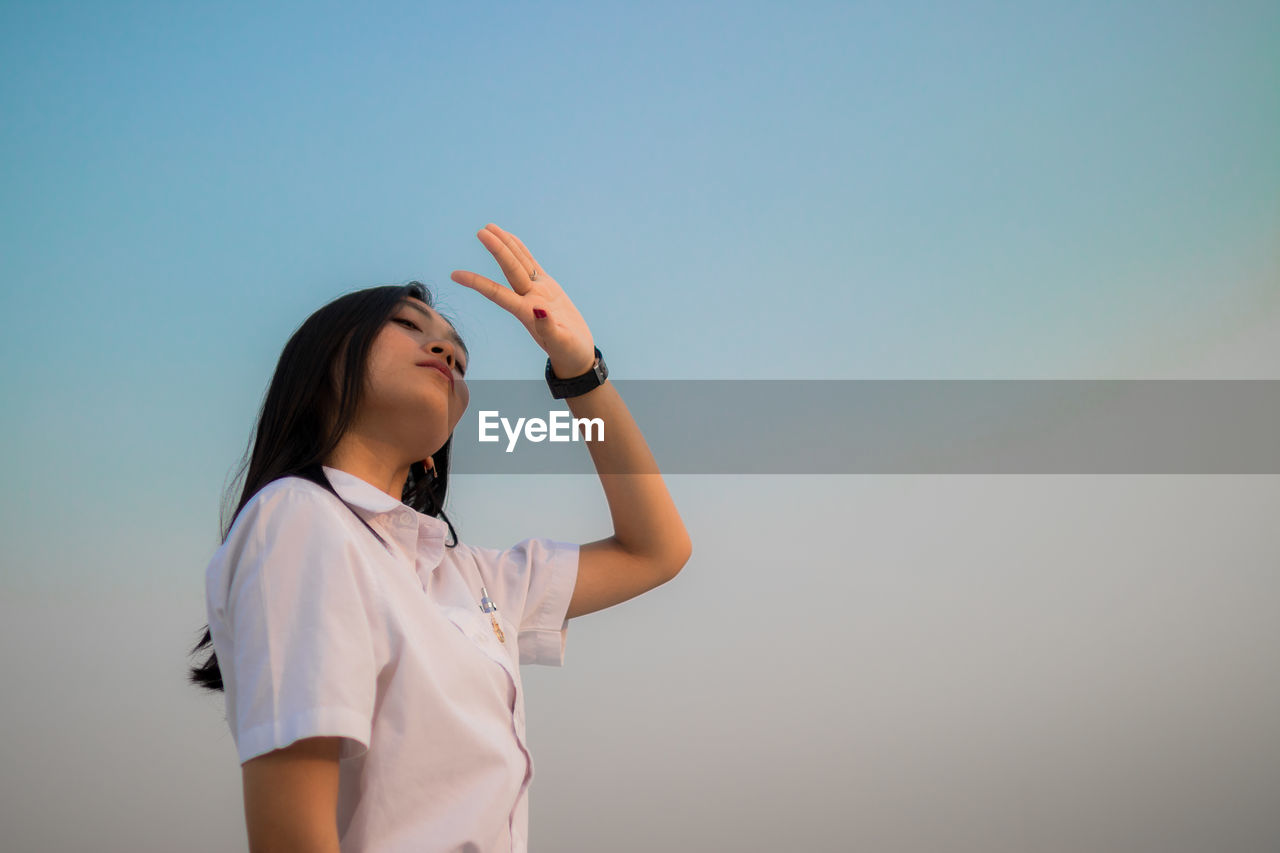 Low angle view of young woman shielding eyes standing against sky during sunset