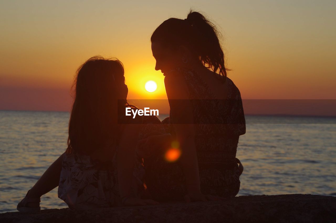 Rear view of woman and girl sitting on beach during sunset