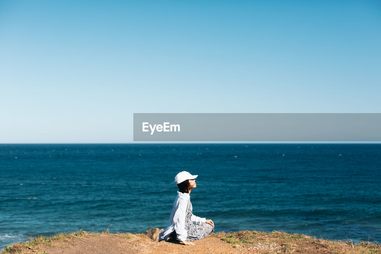 Side view of woman sitting on cliff by sea against clear sky