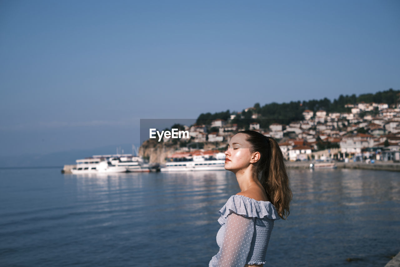 Smiling young woman standing by lake against sky in ohrid city