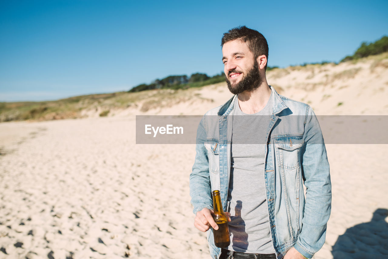 Young man holding alcoholic drink bottle while standing at beach during sunny day