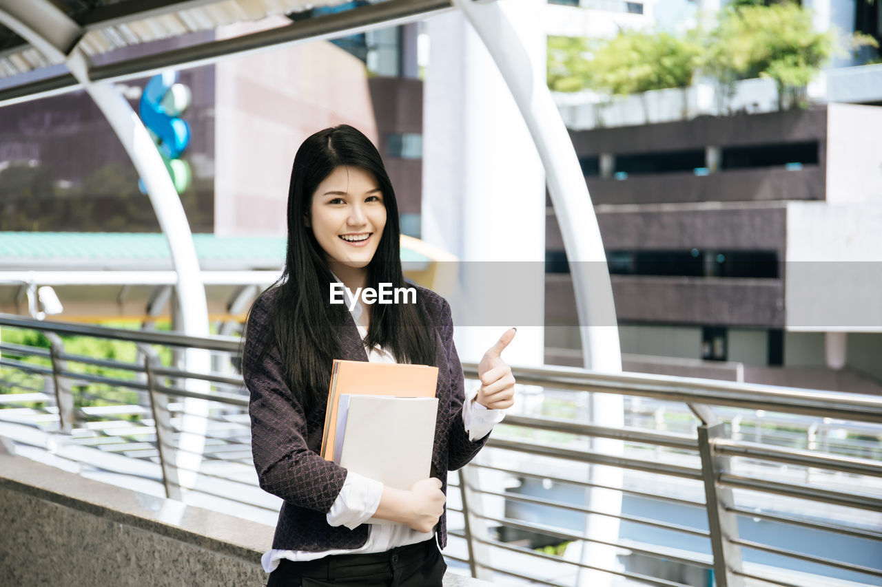 Portrait of woman holding book while standing by railing