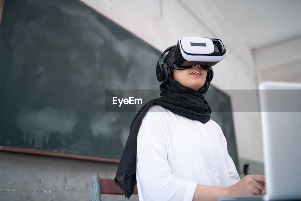 Low angle view of young woman looking through virtual reality simulator in classroom