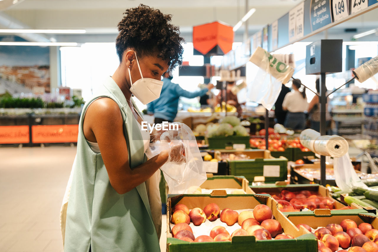 Side view of african american female customer in protective mask and gloves picking fruit from box while making purchases in supermarket during coronavirus pandemic