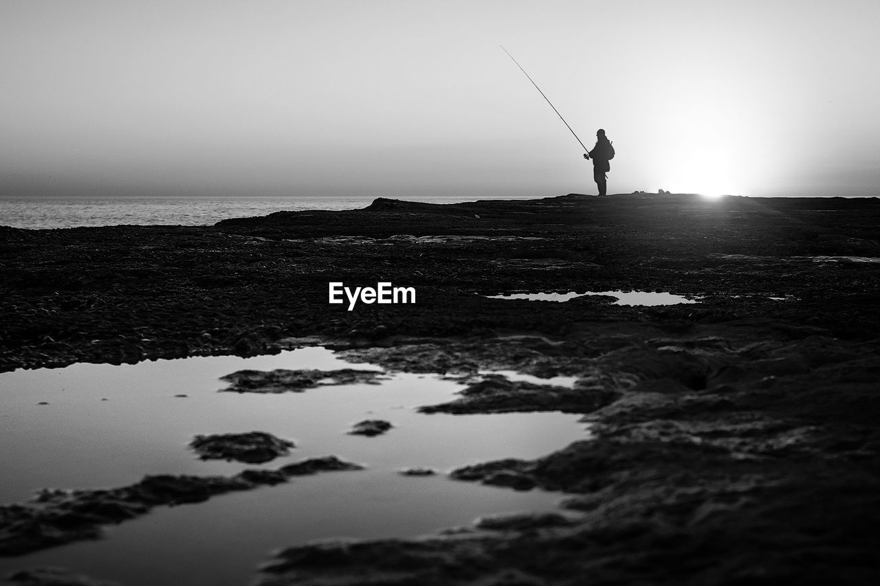 Silhouette fisherman fishing at sea shore against sky during sunset