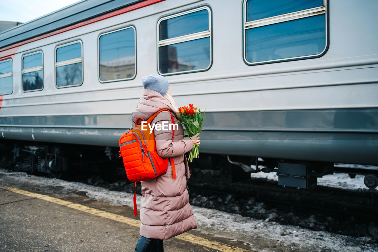 Girl on the platform of the station with a red backpack and a bouquet of tulip flowers