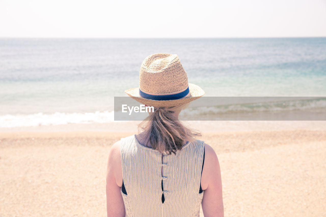 Rear view of mature woman wearing hat while standing at beach against clear sky during sunny day