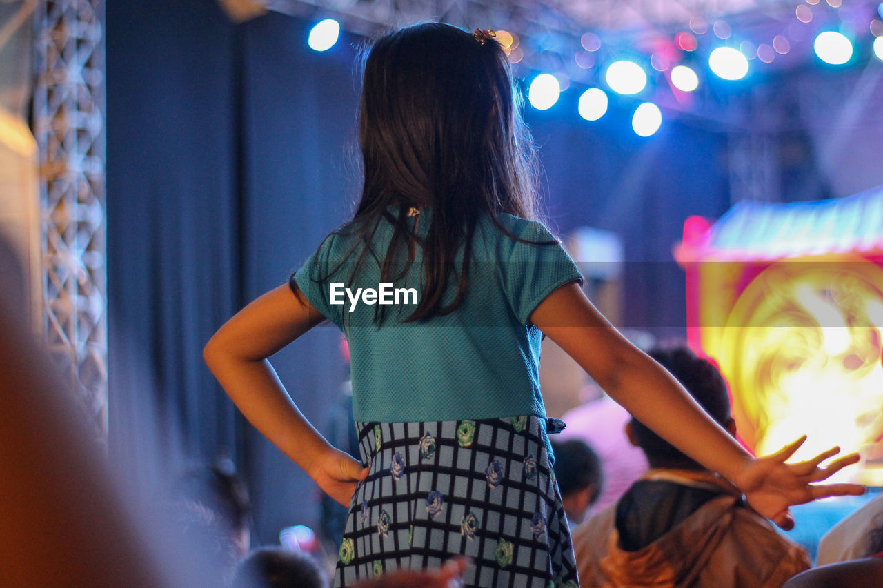 REAR VIEW OF WOMAN WITH ARMS RAISED STANDING IN ILLUMINATED NIGHTCLUB