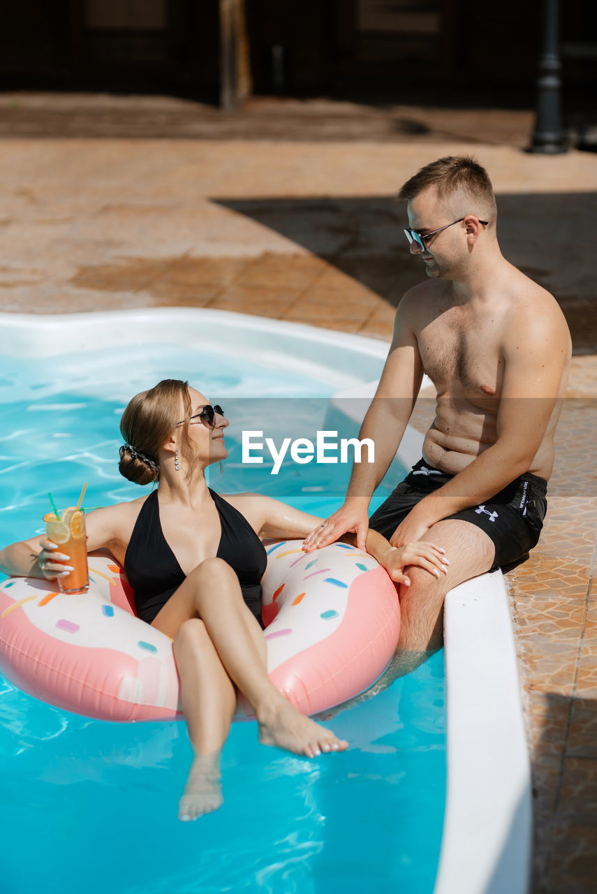 swimming pool, adult, water, two people, swimwear, swimming, women, summer, togetherness, men, relaxation, poolside, young adult, vacation, trip, leisure activity, holiday, lifestyles, nature, female, clothing, happiness, smiling, emotion, enjoyment, positive emotion, friendship, fun, sitting, bonding, bikini, cheerful, sunlight, love, sports, outdoors, day, tourist resort, food and drink, recreation, wellbeing, water park, floating, refreshment, travel destinations, travel, full length, floating on water, child