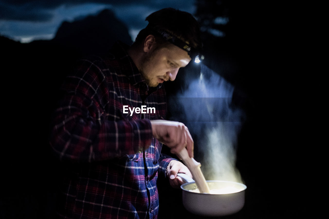 Young man preparing food using headlamp in forest at night