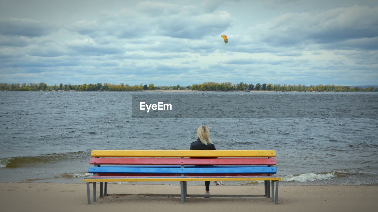 Rear view of woman looking at lake while sitting on colorful bench against sky
