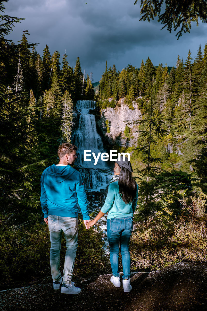 Rear view of people standing against waterfall and trees in forest