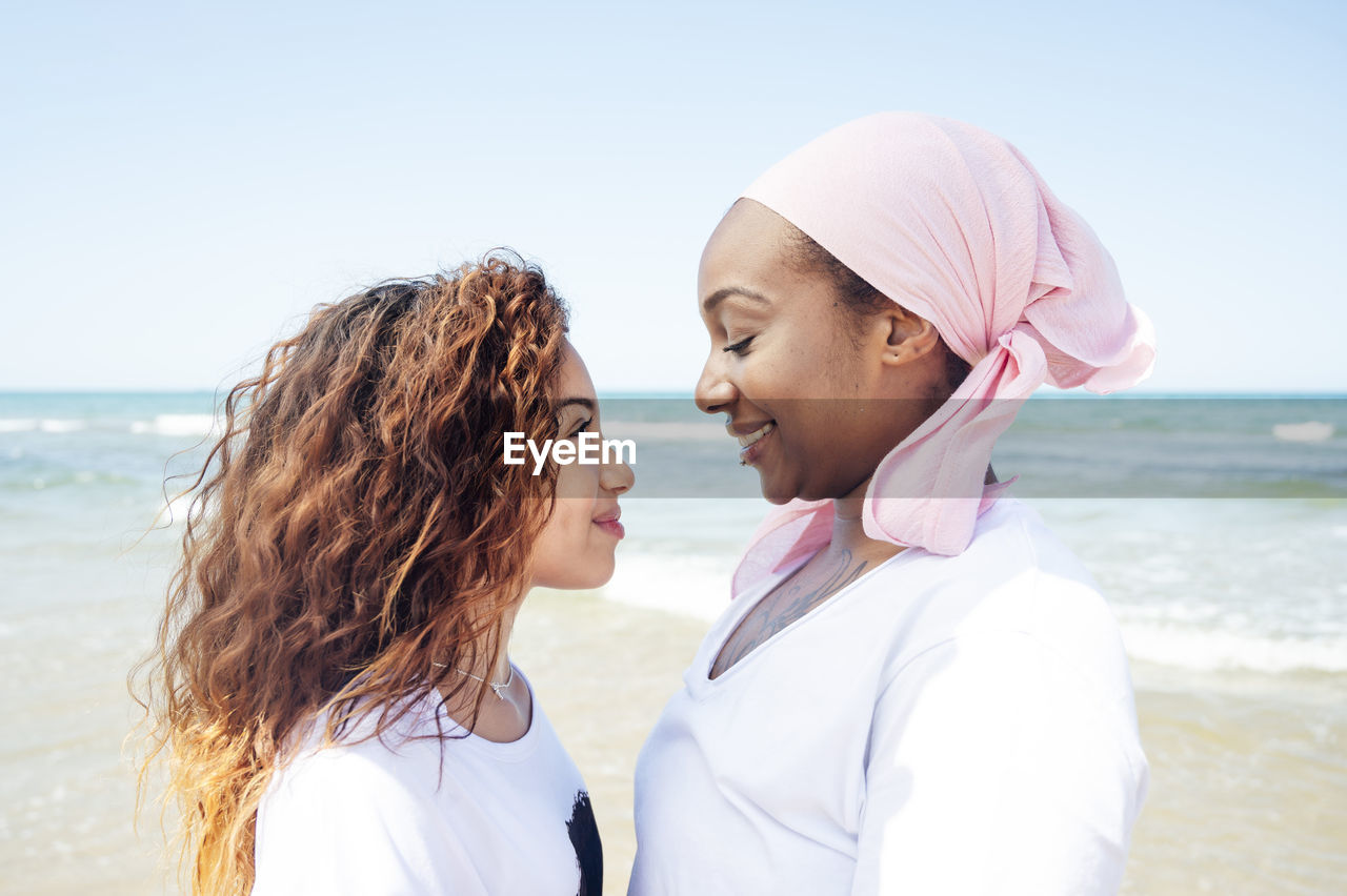 Side view of content african american adult mother and young daughter standing on beach near sea and looking at each other