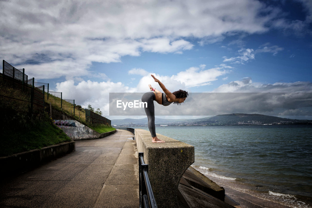 Side view of woman exercising by lake against cloudy sky