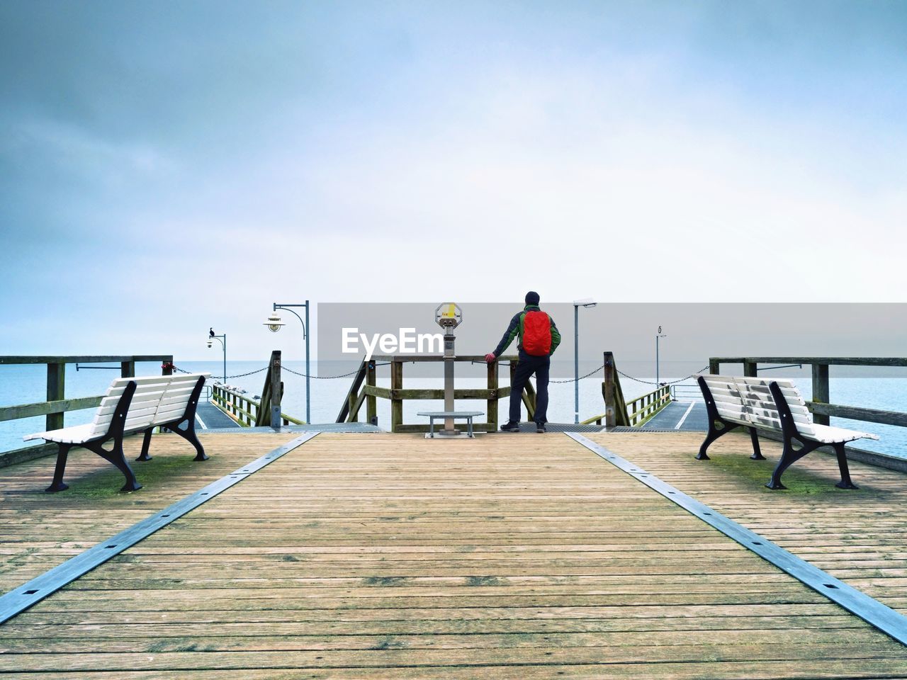 Tourist with red backpack on wooden pier above sea. man in trekking suit in harbor in rainy day