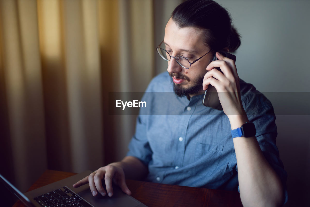 Businessman with a beard and glasses is sitting at a laptop at a wooden table. talking on the phone