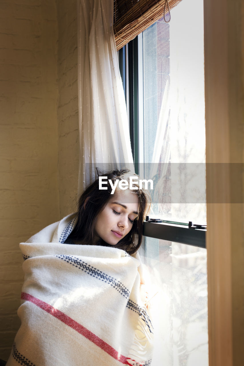 Woman with eyes closed while leaning on window at home