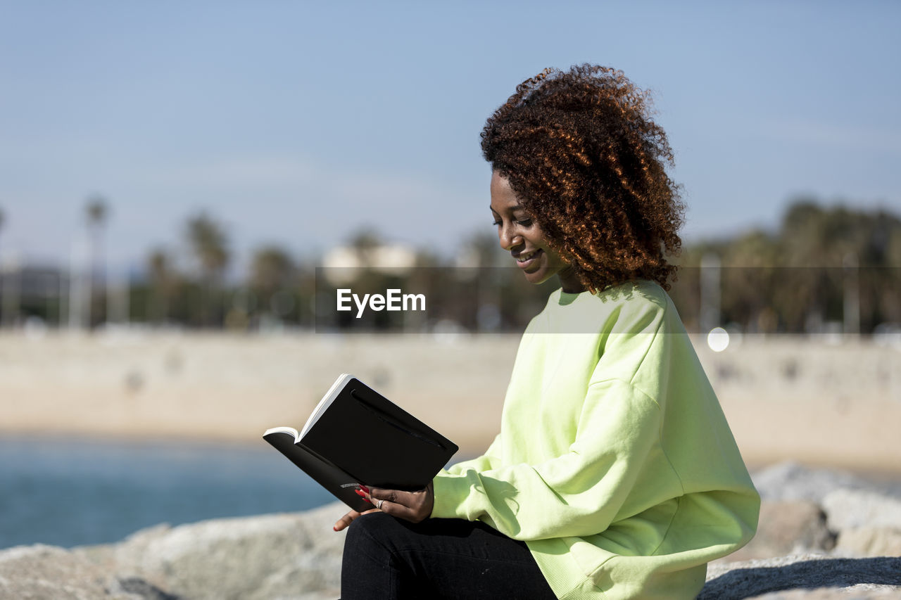 Side view of smiling woman with curly hair reading book while sitting at beach