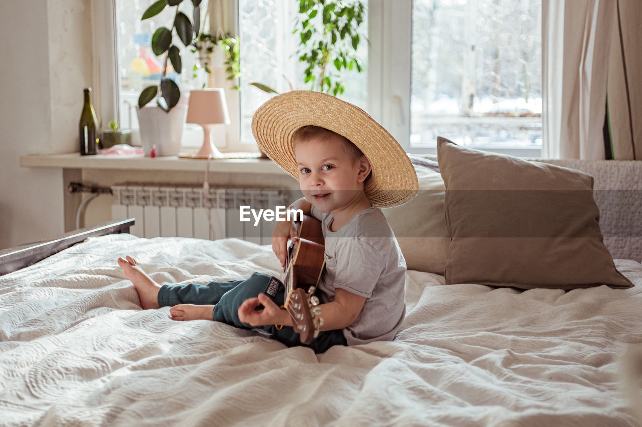 Little toddler boy in hat playing ukulele guitar at home, rustic style. lifestyle concept