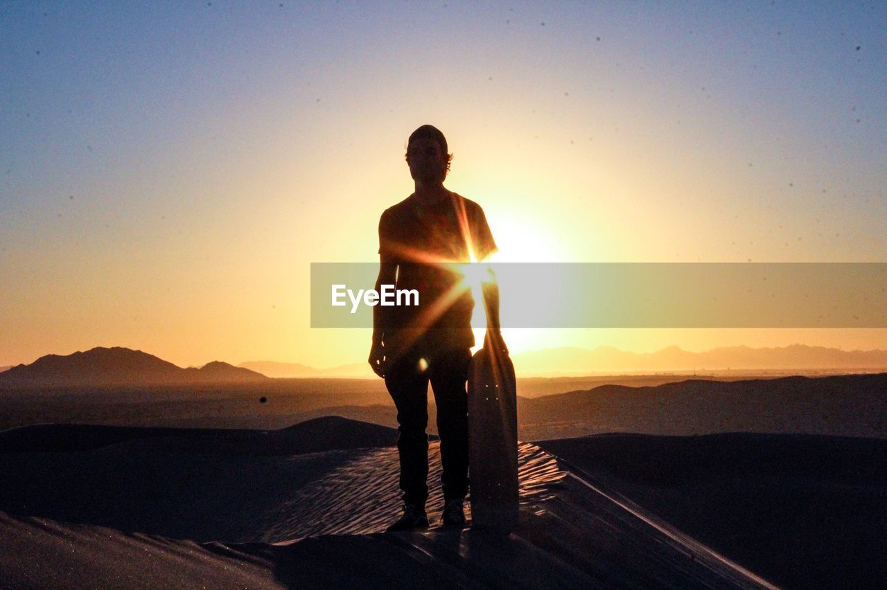 Silhouette man standing with sandboard in desert during sunset