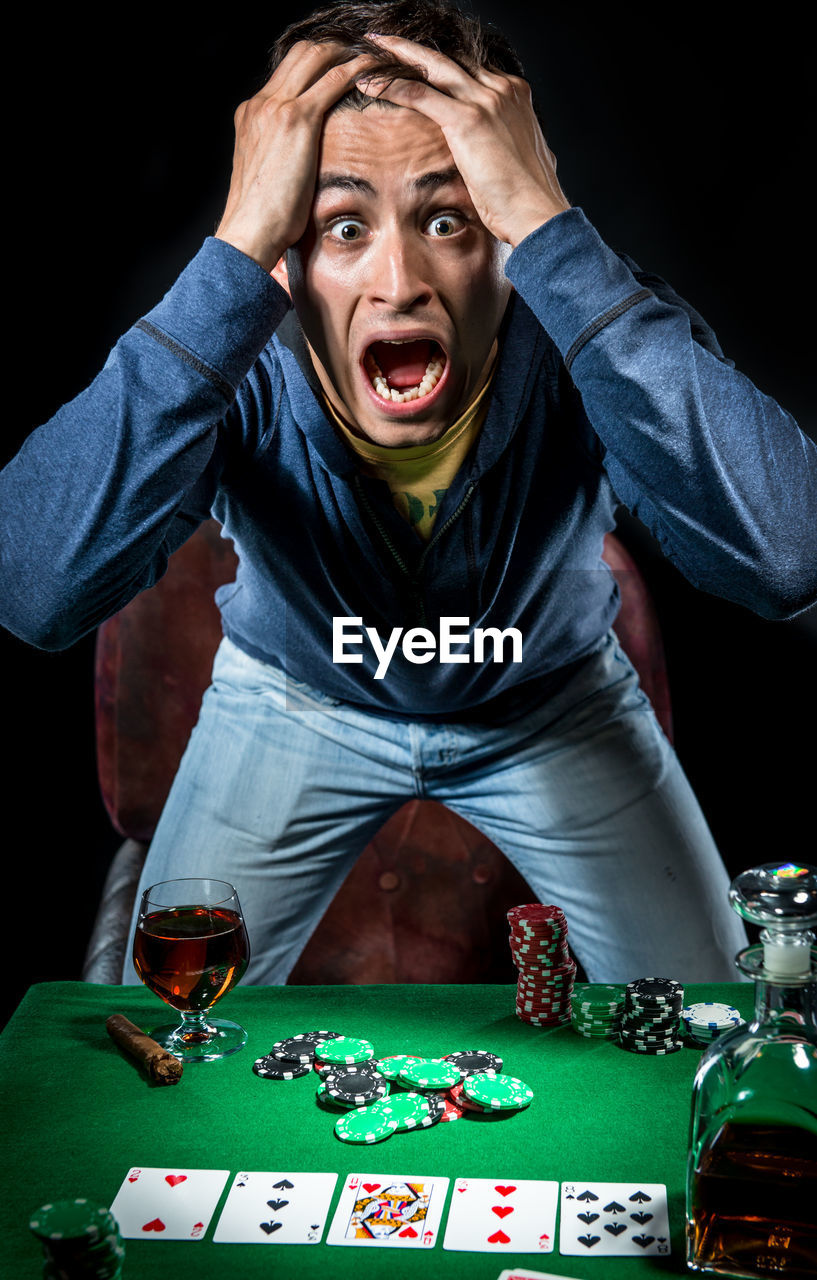 Portrait of shocked man standing at poker table with whiskey against black background