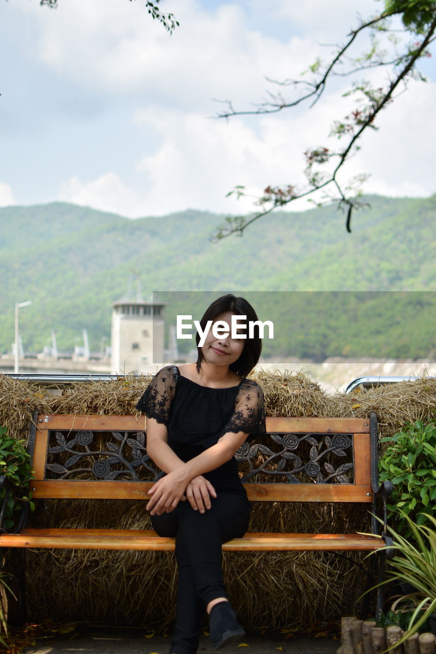 Portrait of smiling young woman sitting on bench against mountains