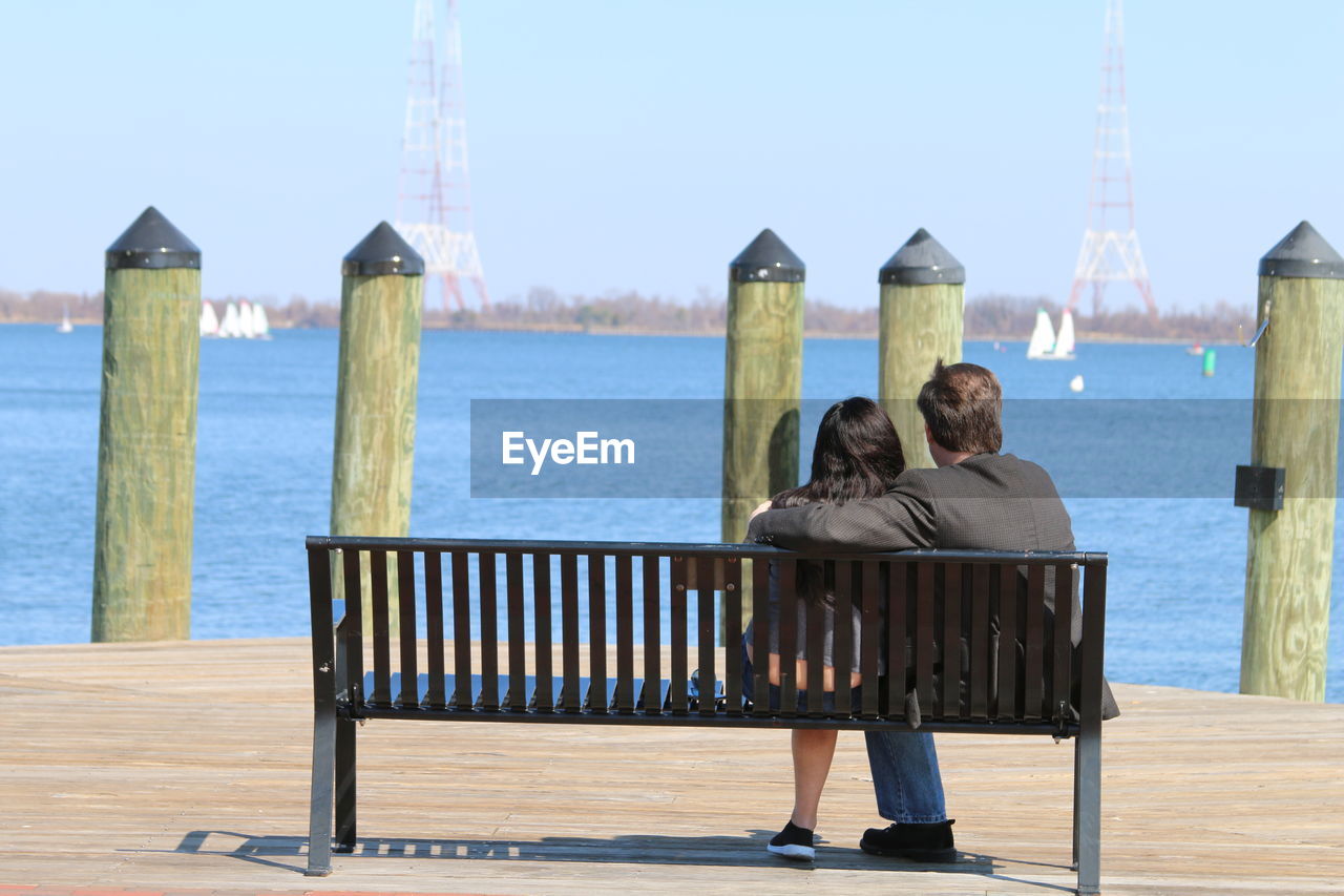 Rear view of friends sitting by sea against clear sky