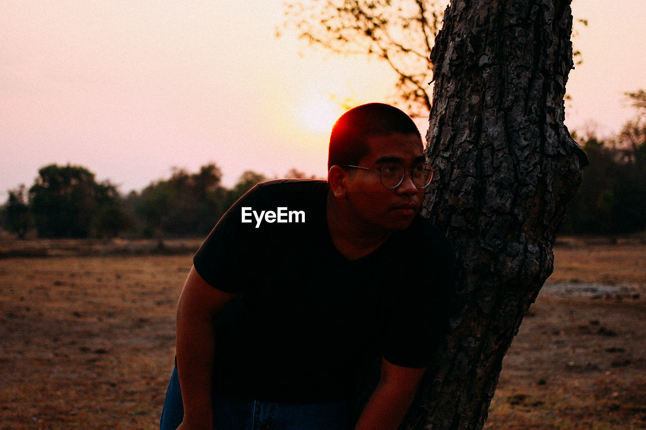 PORTRAIT OF YOUNG MAN STANDING BY TREE TRUNK