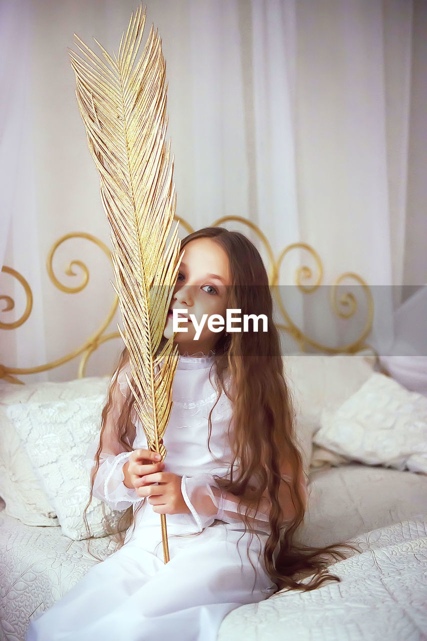Portrait of girl holding golden feather while sitting on bed
