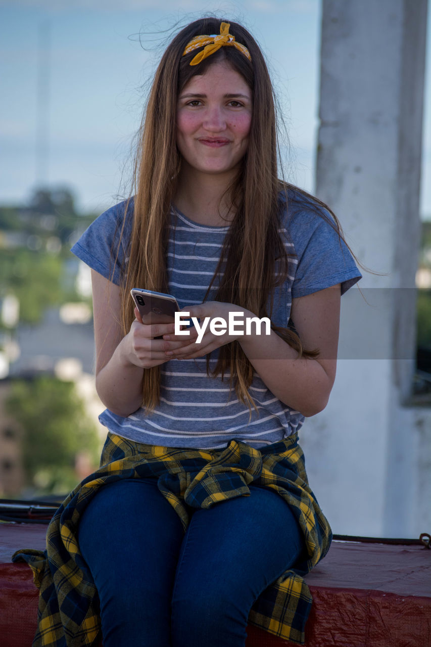 Portrait of smiling young woman using mobile phone while sitting on retaining wall