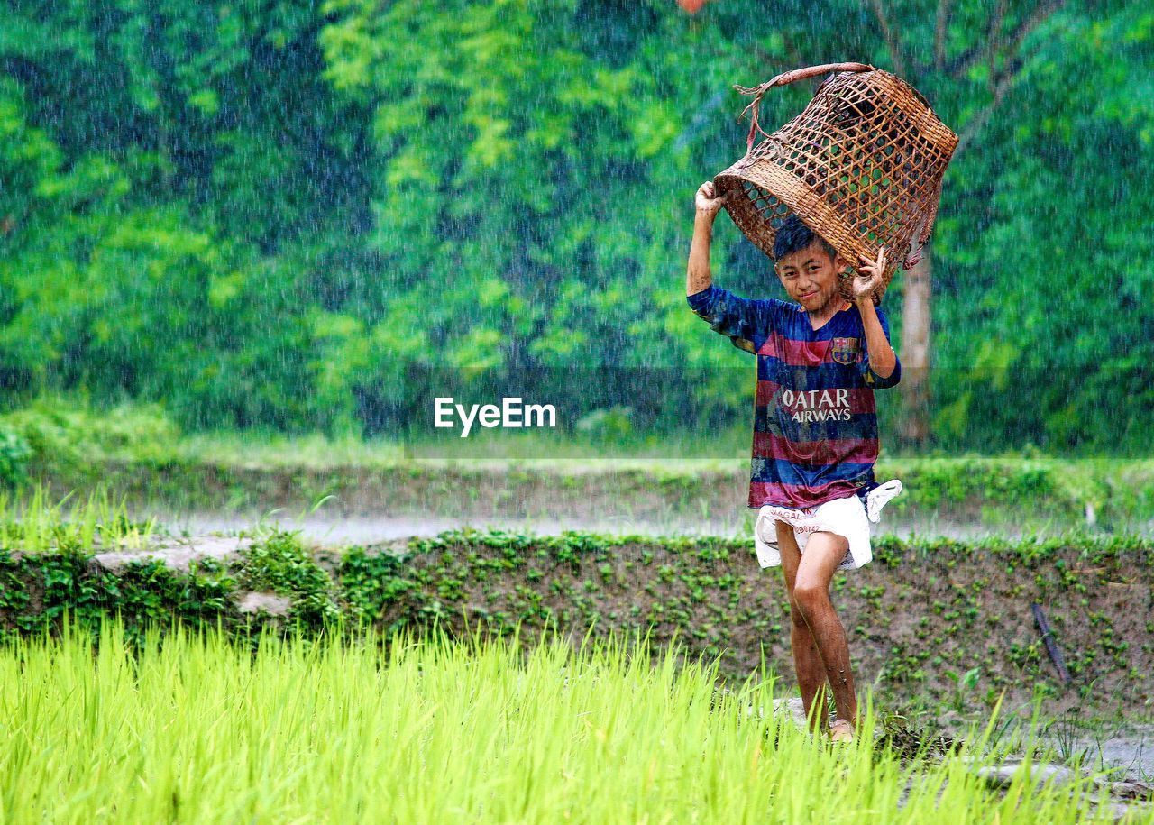 Portrait of boy holding basket while standing on field during rainfall
