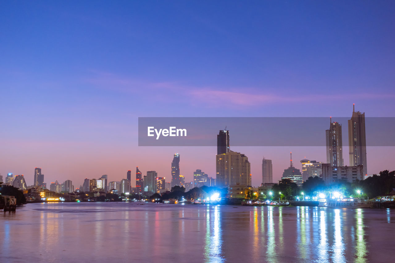 ILLUMINATED BUILDINGS BY RIVER AGAINST SKY IN CITY AT WATERFRONT