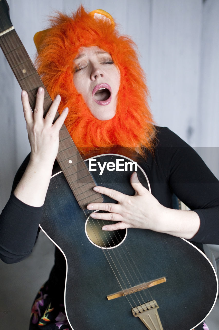 Middle-aged woman wearing lion wig, singing song with her mouth open and playing the guitar