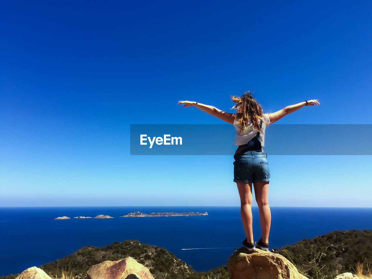 MAN STANDING ON ROCK BY SEA AGAINST BLUE SKY