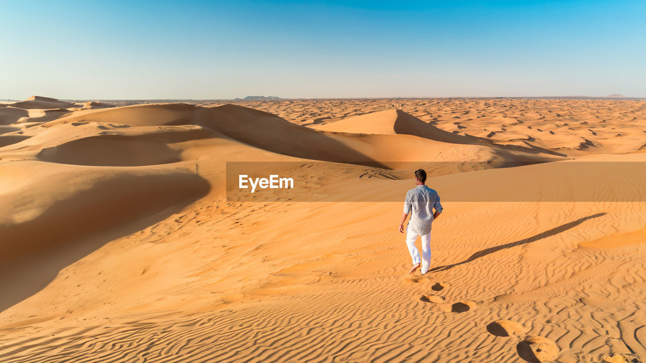 rear view of man walking on sand dune in desert against clear sky