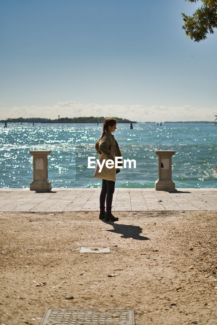 sea, water, vacation, full length, beach, shore, ocean, sky, one person, land, body of water, nature, standing, sand, coast, horizon over water, adult, horizon, leisure activity, casual clothing, day, sunlight, rear view, beauty in nature, holiday, trip, scenics - nature, clear sky, women, sunny, lifestyles, blue, tranquility, outdoors, clothing, young adult, travel, person, tranquil scene, travel destinations, relaxation, looking, solitude, copy space, female, motion, shadow, summer, idyllic